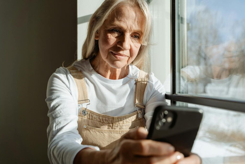 A senior woman enjoys leisure time browsing on her smartphone by a sunlit window. Learn how to make font size larger on any device.